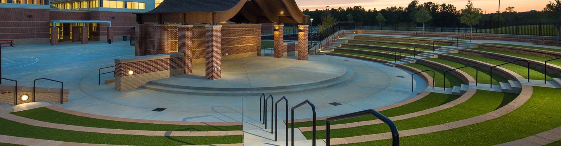 Southwest Greens synthetic grass installation lining tiered amphitheater seating at a brick school or civic building at dusk.